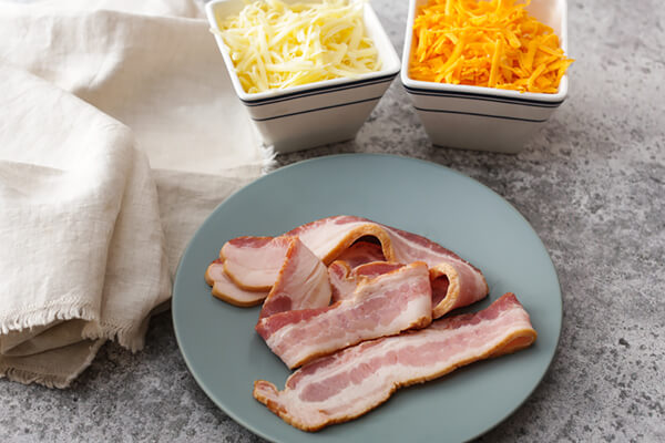 Cheese in bowls and raw bacon on a plate.