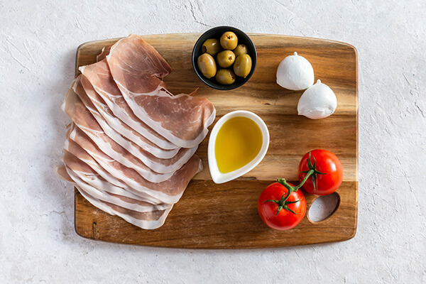 Prosciutto, olives, eggs, and tomatoes on a cutting board.